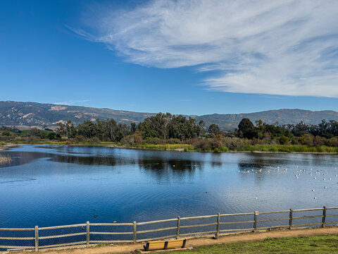 Goleta, CA, USA - December 27, 2023: Lake Los Carneros Park. Dam of reservoir is viewpoint looking N to Santa Inez mountain range. Birds and ducks on water. Green trees and blue cloudscape