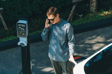 Young man travel with EV electric car charging in green sustainable city outdoor garden in summer...