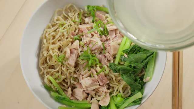Hand pours bowl of clear broth over Bakmi wheat based noodles with chopsticks on side