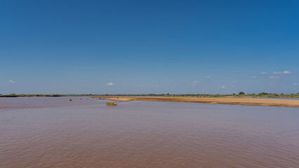 Fototapeta na wymiar A wide calm river with red-brown water. A man in a boat loaded with hay is floating in a riverbed. Tiny figures of people washing clothes are visible. Clear blue sky. Copy space. Madagascar.