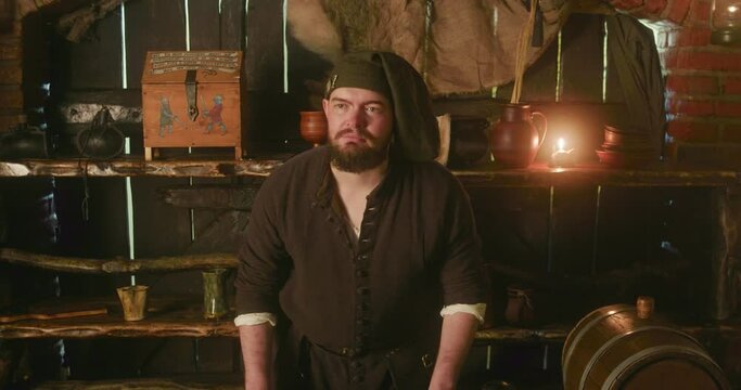 Bearded keeper in bonnet waits for guests in tavern. Stubby dwarf innkeeper stands by counter against rustic wooden shelves in store. Fantasy character