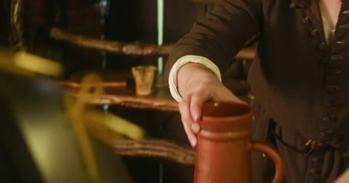 Medieval bartender pours drink at counter closeup. Barkeeper fills clay jug with alcohol from large mug working in tavern. Guest treating in pub