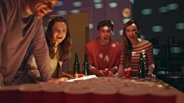 Friends playing beer pong at home party with flashing lights. Man throwing ball