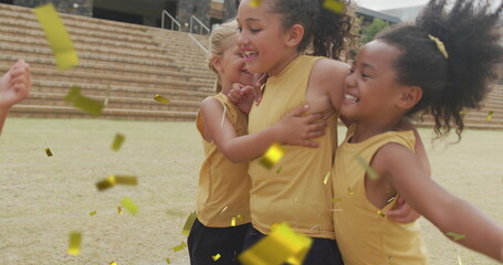 Image of gold confetti over happy diverse schoolgirls celebrating and hugging on sports field