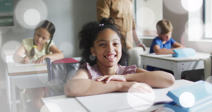Image of white bokeh light spots over smiling biracial girl at desk in diverse class