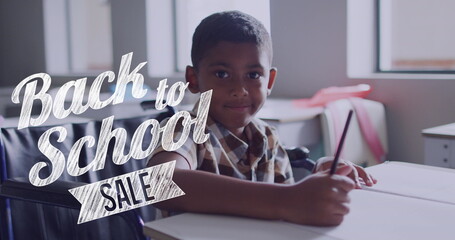 Image of back to school sale text over smiling biracial schoolboy working at desk in class