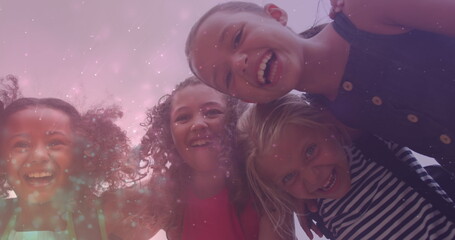 Image of light particles over happy diverse schoolgirls embracing outdoors