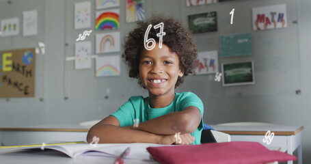 Image of numbers over happy biracial schoolboy sitting at desk in class smiling