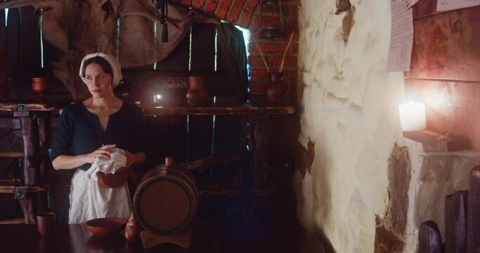 Worried waitress wipes forehead in tavern. Medieval maid in apron cleans clay bowl standing at counter in guesthouse. Busy woman works in inn