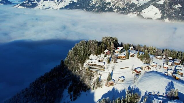 Aerial view of a small Swiss village nestled in the clouds. The village is covered in snow and surrounded by mountains. The view is stunning and otherworldly.
