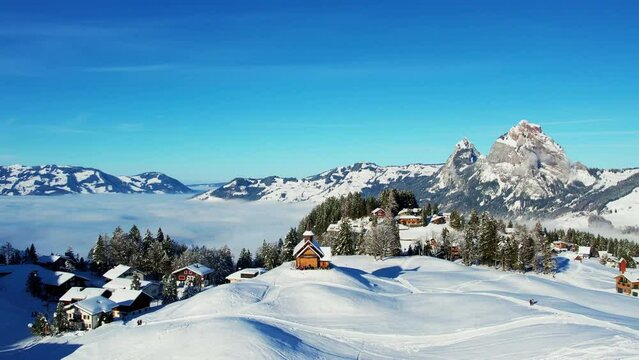 Aerial view of the snow-covered Swiss Alps, with the village of Stoos nestled in the valley below. The mountains are covered in a blanket of white snow.