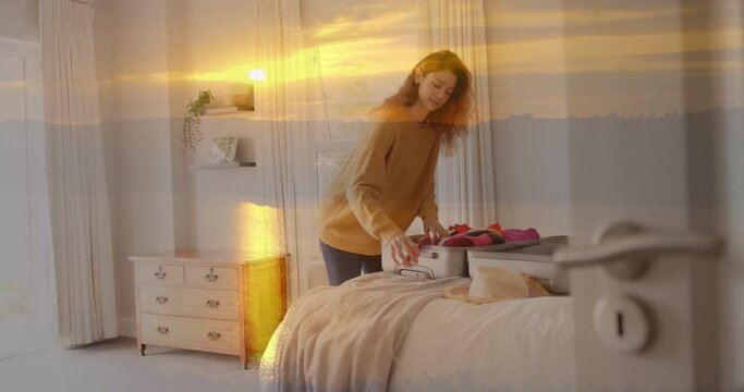 Animation of happy caucasian woman packing suitcase in bedroom over sunset sky