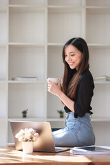 Smiling woman enjoying tea and coffee in her kitchen at home, sitting indoors with a laptop, phone, and a book, radiating happiness and casual beauty