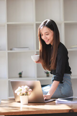 Smiling woman works on her laptop at home in the kitchen, surrounded by coffee, a book, and technology on a cozy interior table