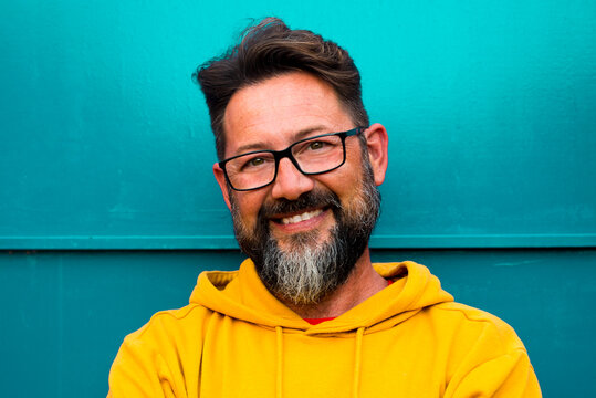 Portrait Of Cheerful Adult Handsome Man Smile With Yellow Sweater Clothes And Blue Wall In Backgorund - Happy People And Copy Space - Beard 50 Years Old Male With Black Eye Glasses