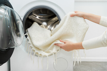 Woman unloading laundry, taking white wool blanket out of the drum of washing machine in laundry...