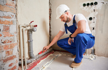 Male plumber in safety construction helmet using spirit level tool while installing pipe system at home. Man measuring pipe level with building instrument in apartment under renovation.