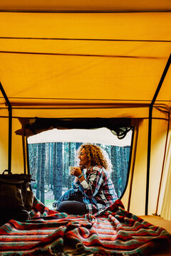 Happy lonely adult caucasian woman sit down outside a tent enjoying a cup of tea and the wild outdoor nature around - free alternative camping vacation with tent and backpack for wanderlust people