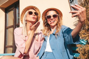 Two young beautiful smiling hipster female in trendy summer clothes. Sexy carefree women posing on street background. Positive models having fun, hugging at sunset. In hat and sunglasses. Take selfie