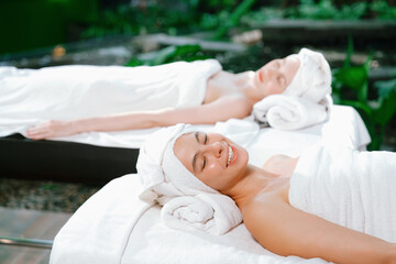 Two beautiful young woman lie on spa bed with white towel while felling in deep relaxation rounded by relaxing and calming nature. Healthy and beauty concept. Blurring background. Tranquility.