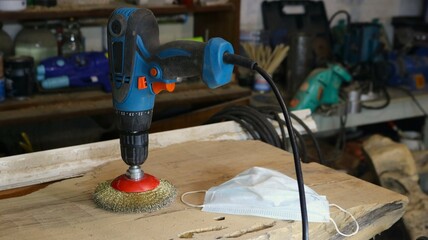 a drill with an attachment made of metal bristles standing on a wooden board in a craftsman's workshop, an apparatus for working with wood and a dust mask as a carpenter's working tools on a workbench