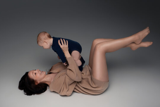 young mother with long dark hair in a beige sweater lies on the floor and plays with her little son. a child and a parent play with each other, photo on a dark background. space for text