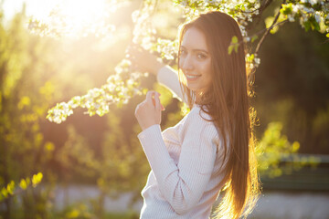 Portrait of a beautiful young woman in a beautiful beige dress smiling with her teeth laughing on a warm spring day at sunset against the backdrop of a green blooming park garden. - obrazy, fototapety, plakaty