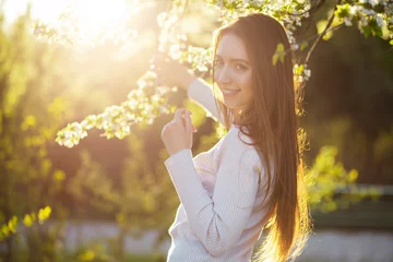 Tuinposter Portrait of a beautiful young woman in a beautiful beige dress smiling with her teeth laughing on a warm spring day at sunset against the backdrop of a green blooming park garden. © Konstantin Zibert