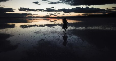 A young woman dance across the mirror like surface of salt lake as the sun disappears behind the clouds.