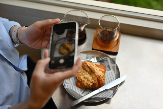Close-up image of Young Asian man using phone to take a picture of croissant and coffee, Focus on croissant