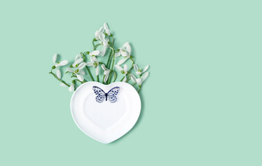 snowdrops flowers, heart plate with decorative butterfly on green background. spring season. hello...