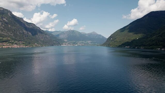 The drone flies over the lake surrounded by green mountains. In the distance you can see the town of Porlezza. Breathtaking scenery on a beautiful summer day