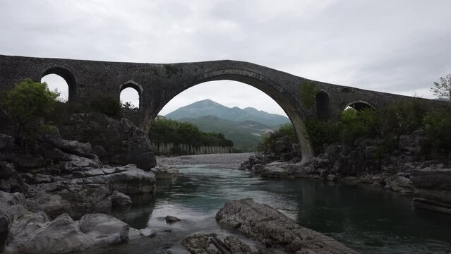 Aerial low angle drone view flying through and under historic stone Mesi bridge in Shkod&euml;r Albania, river view with mountains and cloudy sky during an overcast day