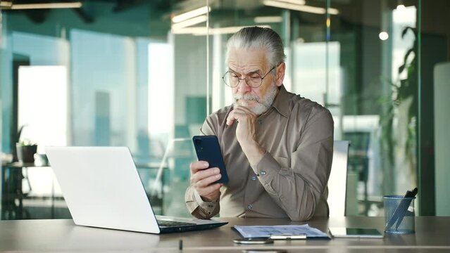 Serious thoughtful senior gray haired bearded businessman is using phone sitting in business office. Focused mature entrepreneur reads message, chats with client, banking in phone app, browsing online