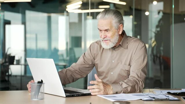 Smiling senior gray haired bearded businessman talking on a video call using a laptop sitting at a workplace in business office. Elderly entrepreneur has a conversation with a friend or colleague