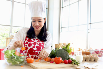 Happy smiling beautiful young Asian woman in red heart apron and chef hat holds sliced carrot for making salad while sits behind kitchen counter. Healthy girl making fresh vegetable salad at kitchen.