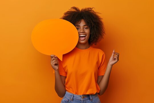 African american woman holding a card in the shape of a big orange speech bubble, orange t-shirt, solid background.