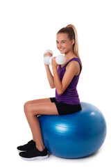 Slim and fit teen girl sitting a swiss blue ball and holding white dumbbells . Full length shot on white background.