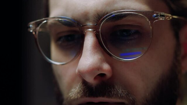 Close-up eyes in glasses of man programmer working at computer at night, light of monitor, binary code reflected in his glasses. Concert programmer in glasses works at computer at night time.