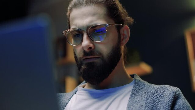Businessman working at night, looking at monitor, meditating with glasses. Remote work. Man looking at smart device and reading social information stream online. Feed screen reflections on glasses.