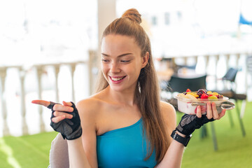 Young pretty sport girl holding a bowl of fruit at outdoors pointing to the side to present a product