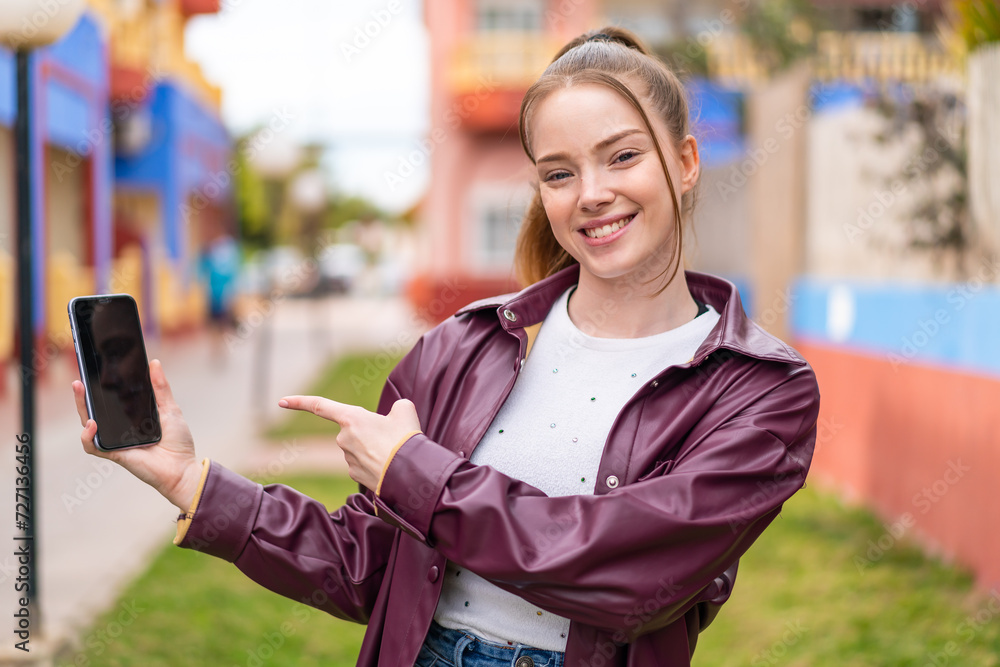 Wall mural Young pretty girl using mobile phone at outdoors and pointing it