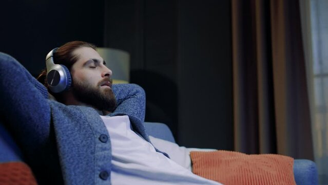 Bearded young man relaxing on comfortable sofa with eyes closed wearing headphones. Handsome guy enjoys listening chill music audio sound meditating feeling no stress at home. Side view.