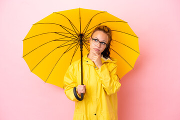 Teenager Russian girl with rainproof coat and umbrella isolated on pink background having doubts