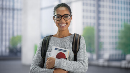 Portrait of a Cheerful African American Student Standing in a Modern Space. Young Black Female Looking at Camera and Smiling Joyfully. Scholar Holding Academic STEM Textbooks with Both Hands