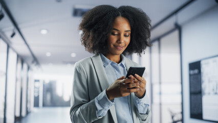 Portrait of a Happy African American Woman Texting on Her Smartphone, Smiling while Standing in a Corporate Office. Young Black Female Marketing Manager Checking Social Network, Chatting with Friends