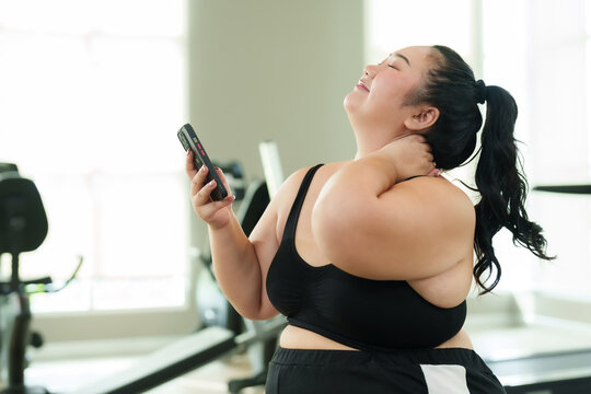 Overweight Asian woman exercise in gym, in black sportswear texting on phone while resting on gym bench. Joyful plus-size female athlete checking smartphone during workout break.