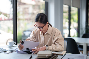Middle aged business woman reading a book and checking financial document to taking notes in paper while analysis investment and financial strategy of new business startup in modern workplace