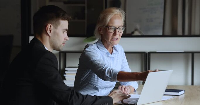 Serious elder and younger colleagues discussing online project at laptop, talking at computer, looking and pointing at screen, sitting at workplace together, cooperating on startup