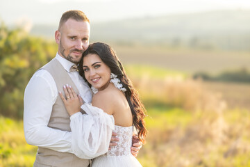 Obraz premium Wedding portrait of smiling newlyweds. A stylish groom in a beige suit and a cute brunette bride in a white dress are tenderly embracing in the park